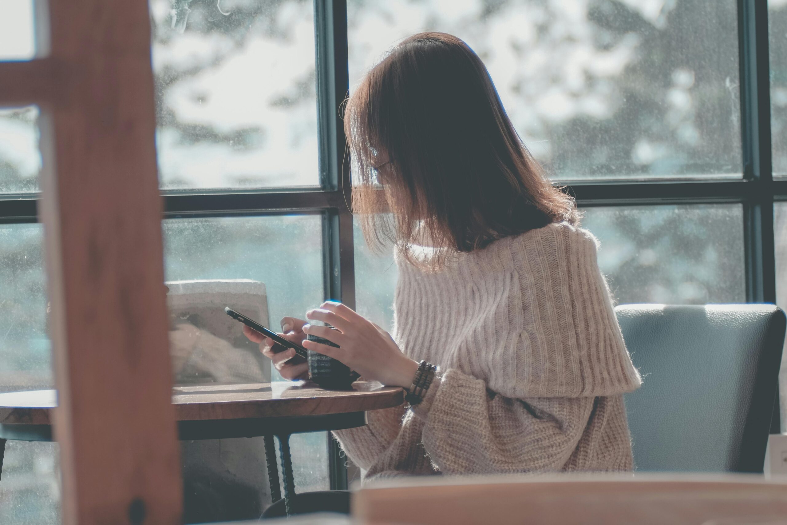 A woman relaxing indoors with coffee, texting on her smartphone by a window.